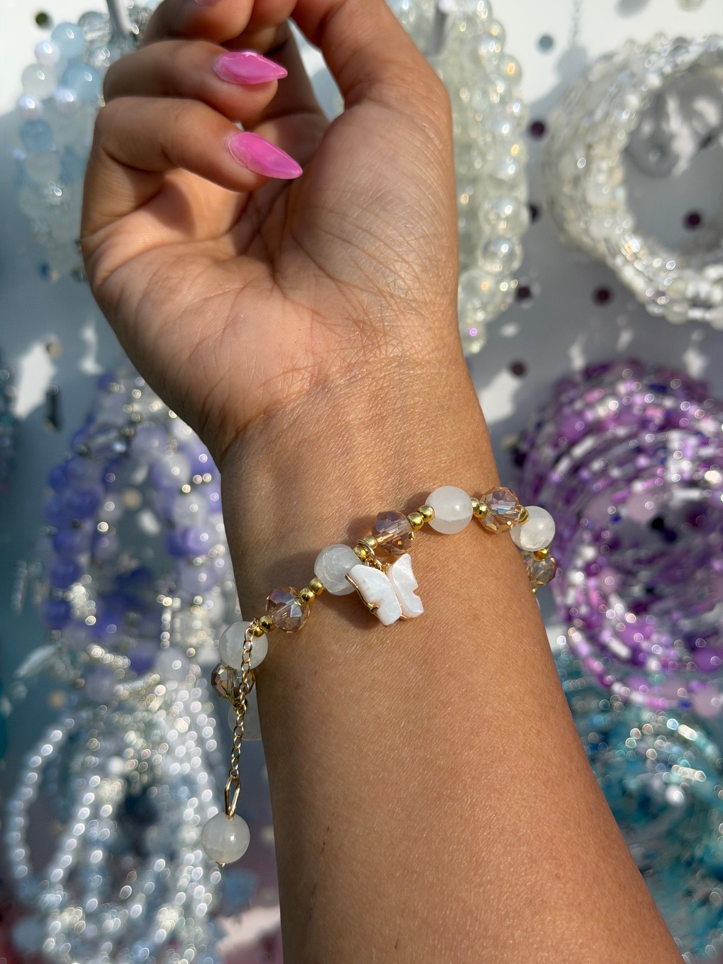 White crystal beaded bracelet with a butterfly charm, featuring gold accents, worn on a wrist with blurred jewelry in the background.