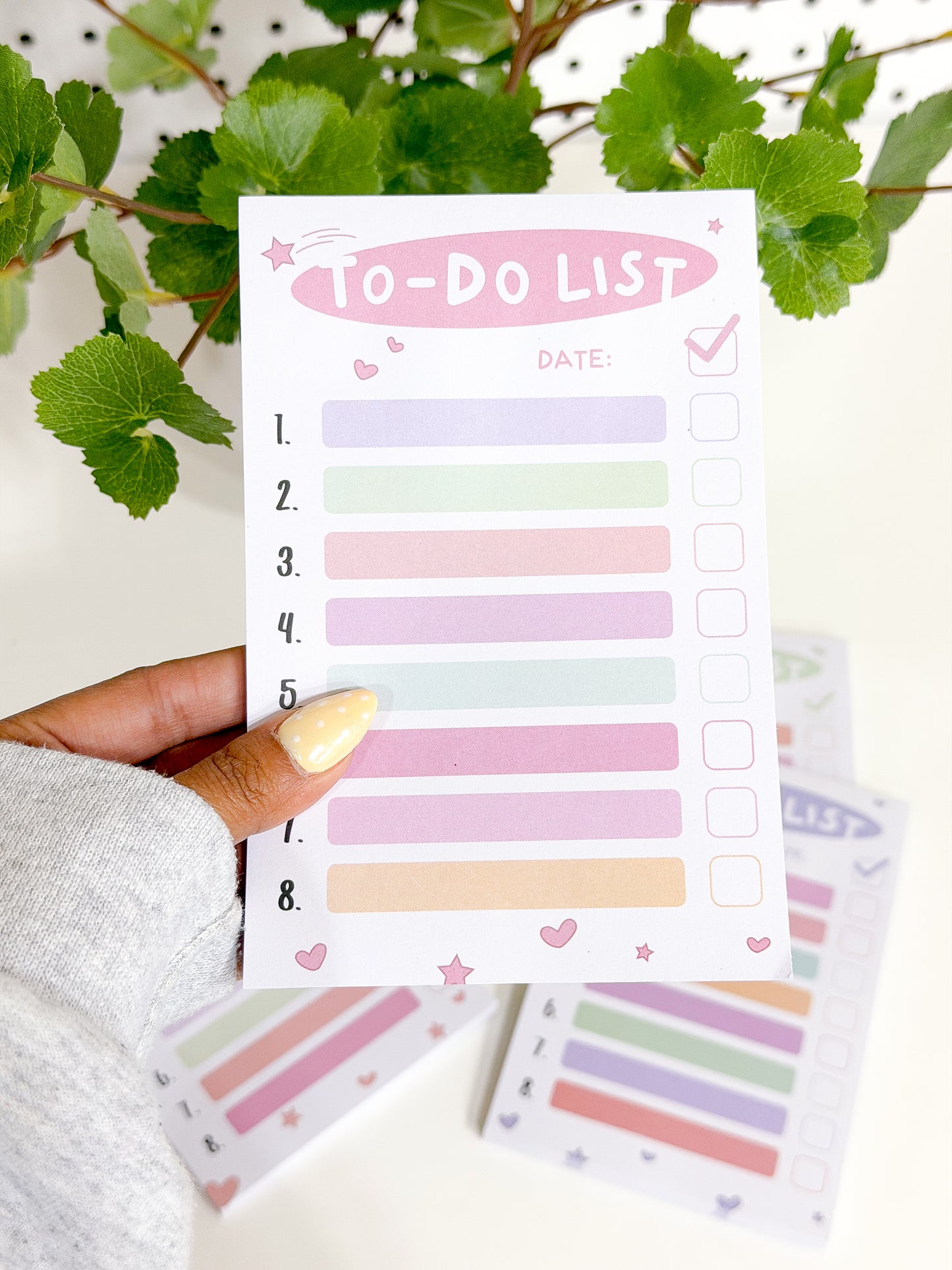 Pastel Tasks Notepad with numbered lines and checkboxes, held by a hand, featuring a 'TO-DO LIST' title and green leaves.