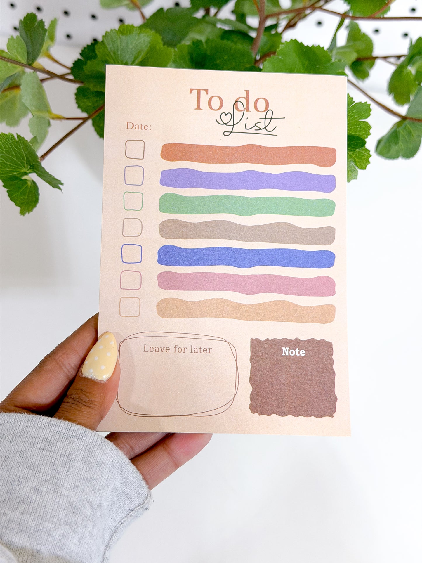 Peach 'To Do List' notepad with colorful wavy lines, held by a hand with polka dot nail polish, against a white background with green foliage.