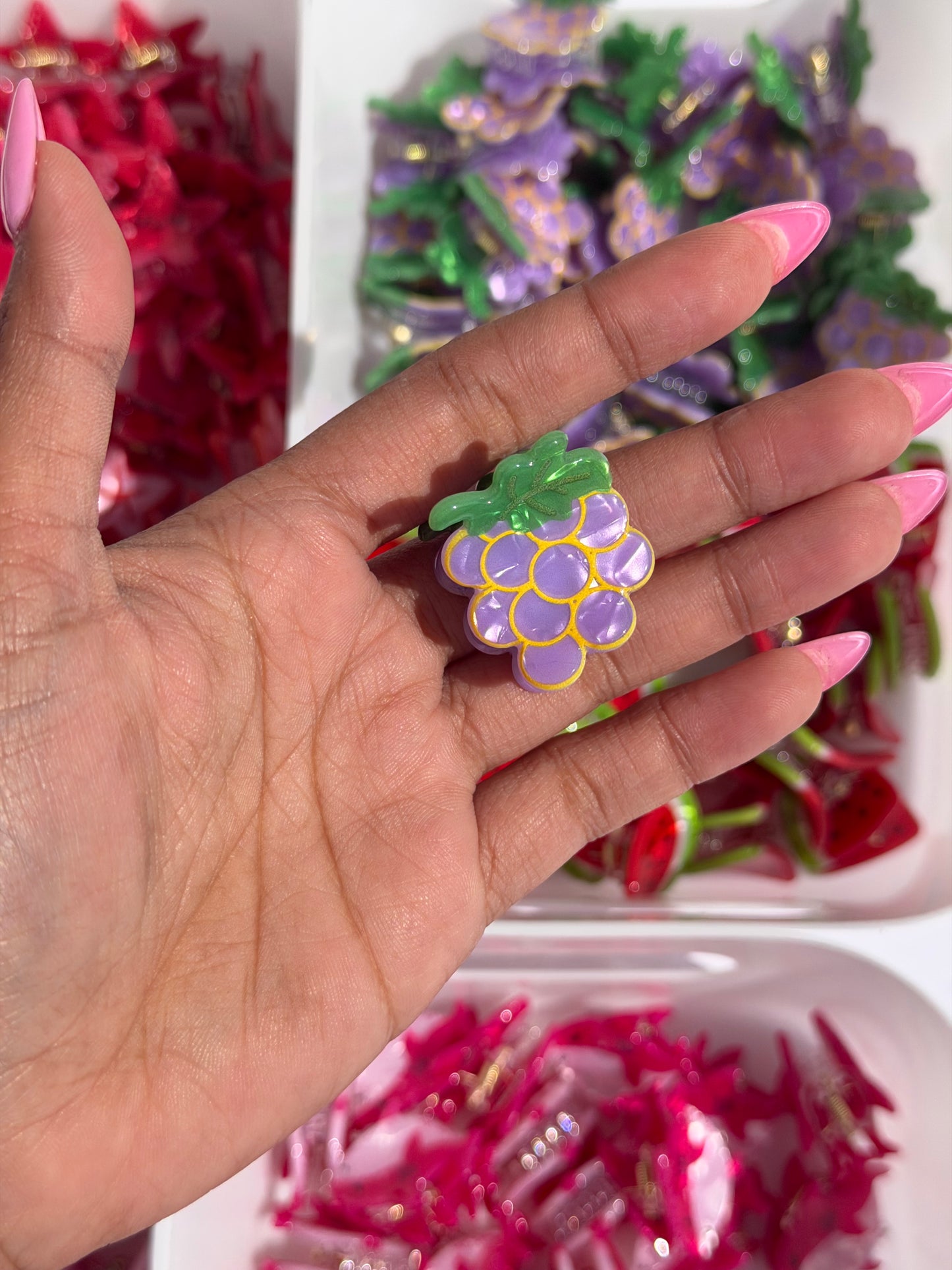 Grape-shaped mini hair clip in purple, green, and yellow, held by a hand with pink nails.