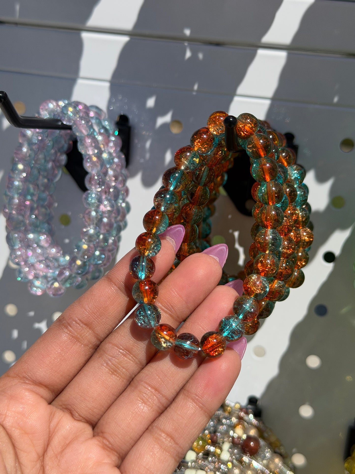 Blue beaded bracelets with crackled blue and orange beads, held by a hand with purple nails, against a white pegboard.