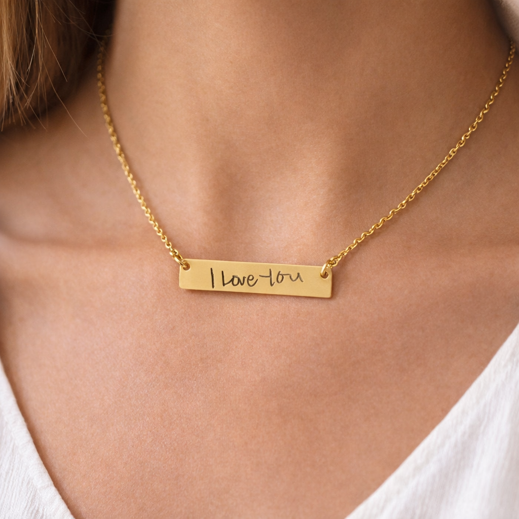 Gold bar necklace with handwriting engraving by brand name, on a model against a light-colored shirt.