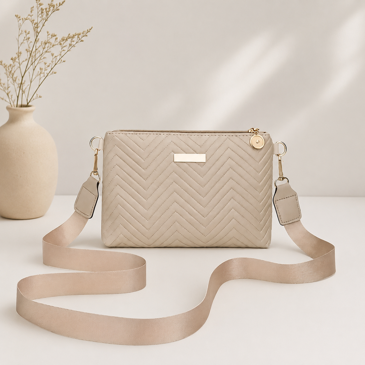 Beige quilted crossbody bag with wide strap and gold-tone hardware, front view on white surface with vase in background