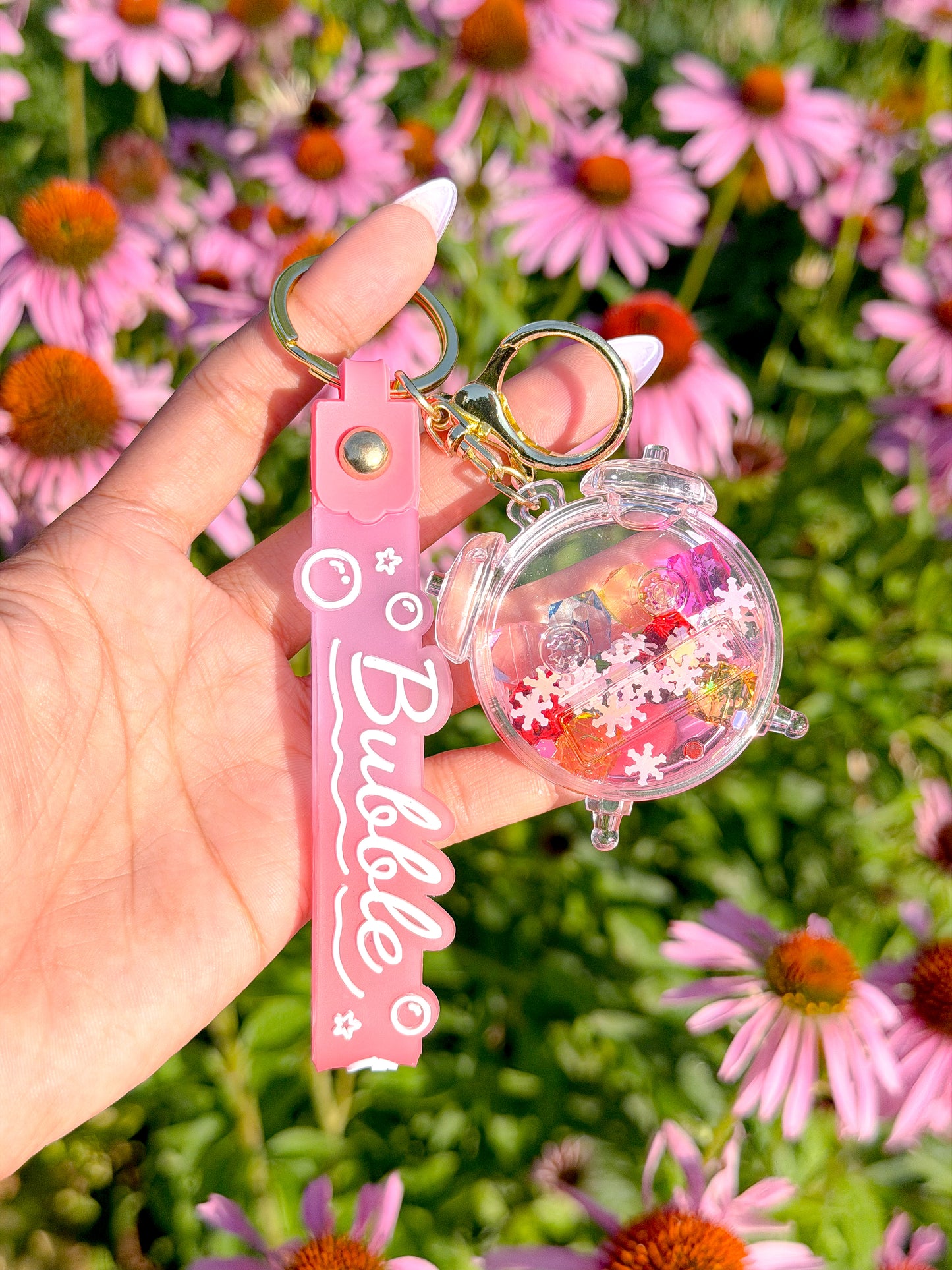 Pink confetti bubble clock bag charm with clear resin snowflake glitter, attached to scalloped tag with "Bubble" text, held against pink flowers.