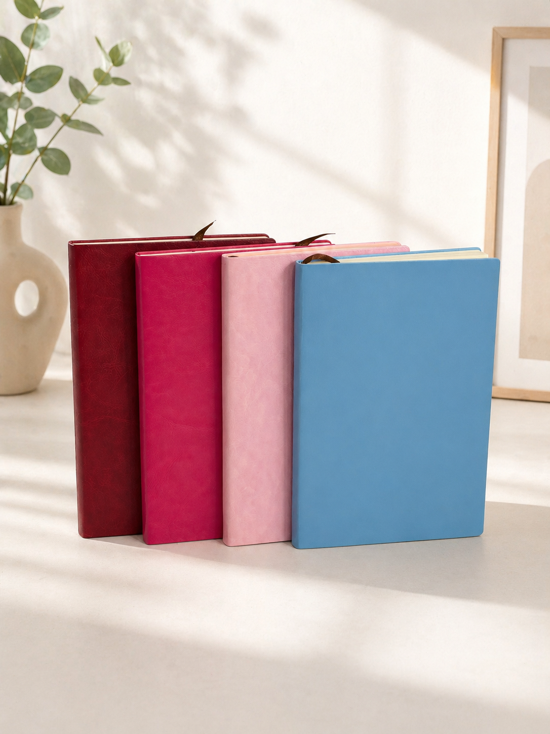 Four A5 size PU leather journals in dark red, fuchsia, light pink, and light blue, standing upright with ribbon bookmarks in a sunlit room.