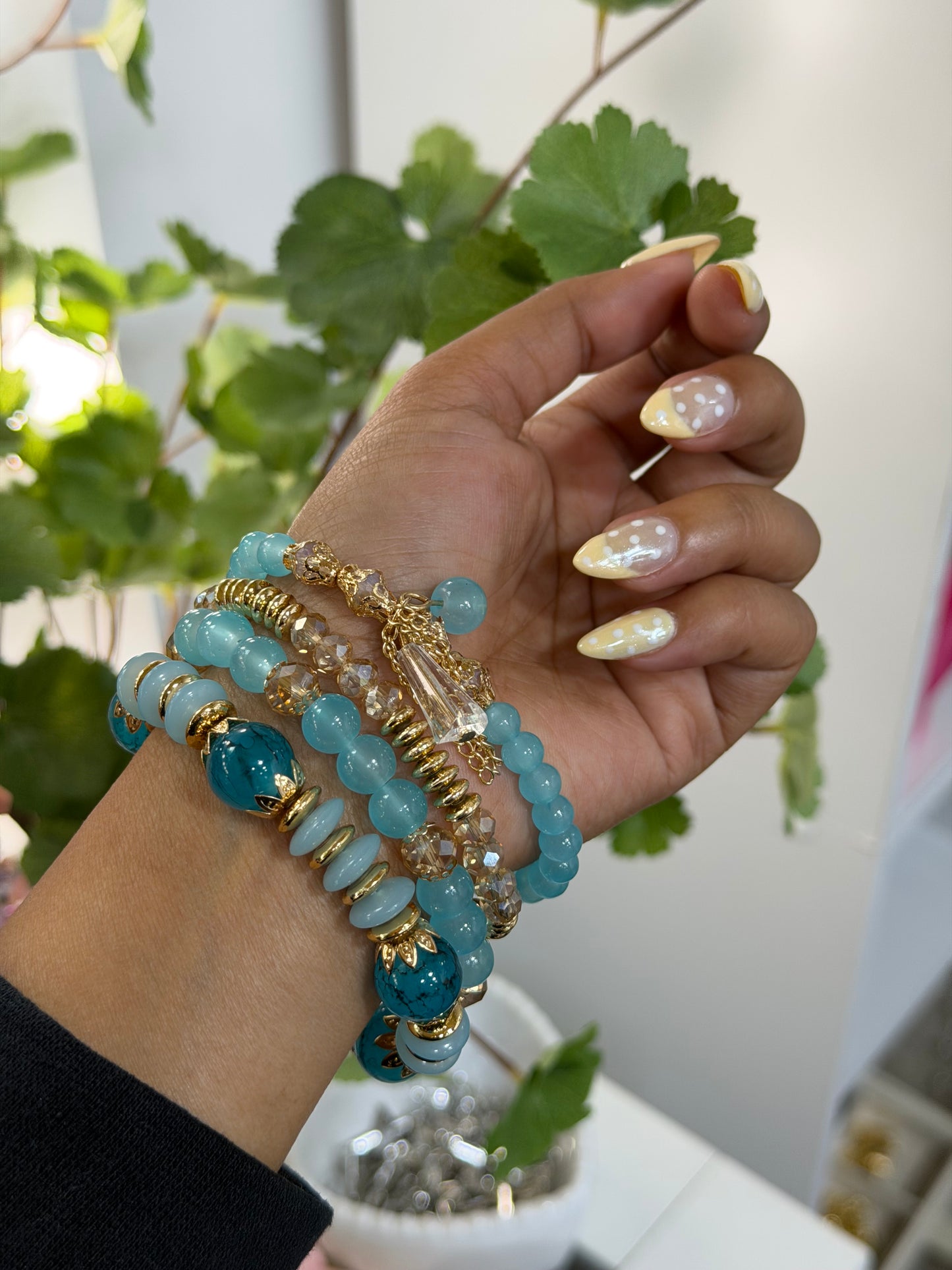 Layered blue and gold beaded charm bracelets with a clear faceted charm, worn on a hand with yellow polka dot nails.