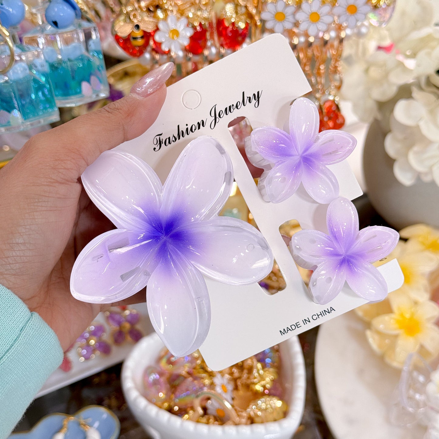 Purple plumeria hair clip sets from Fashion Jewelry, displayed on a product card in a hand.