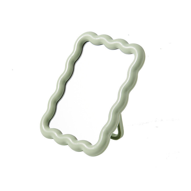 Light sage green ripple tabletop mirror with a wavy-edge frame and kickstand, angled view.