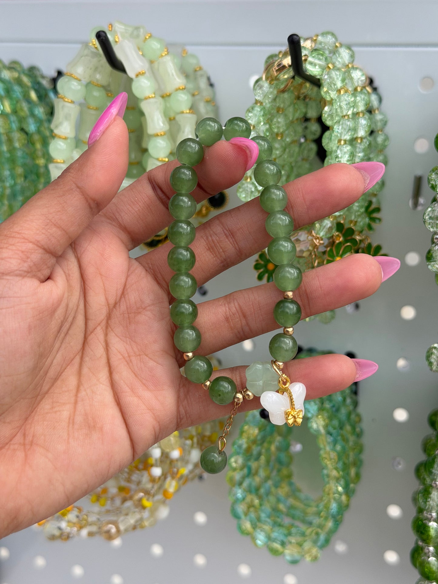 Green beaded bracelet with round beads, gold spacers, and a white butterfly charm, held by a hand with pink nails.
