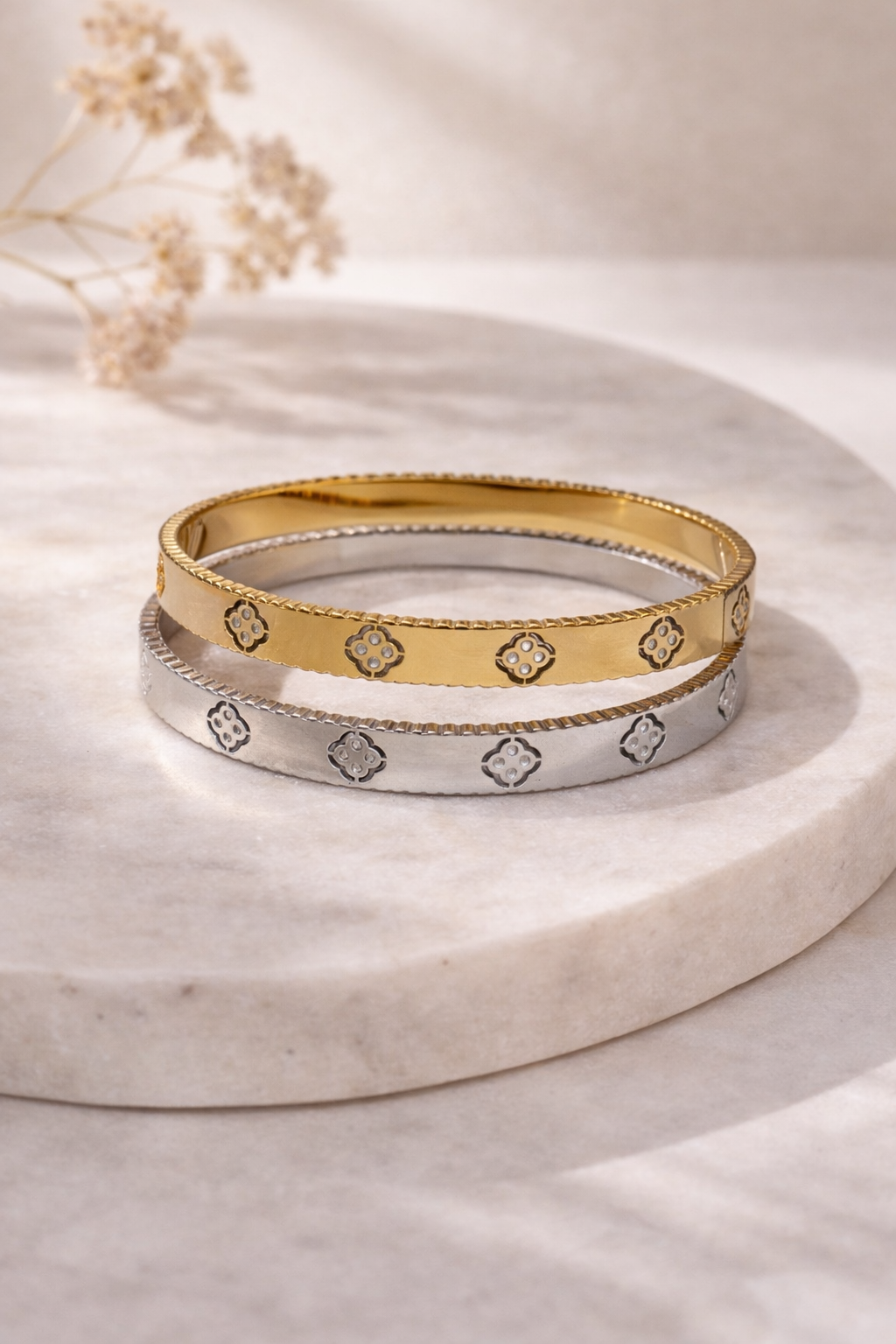 Two stacked bangle bracelets, one gold and one silver, both featuring textured edges and clover cut-out patterns, displayed on marble.