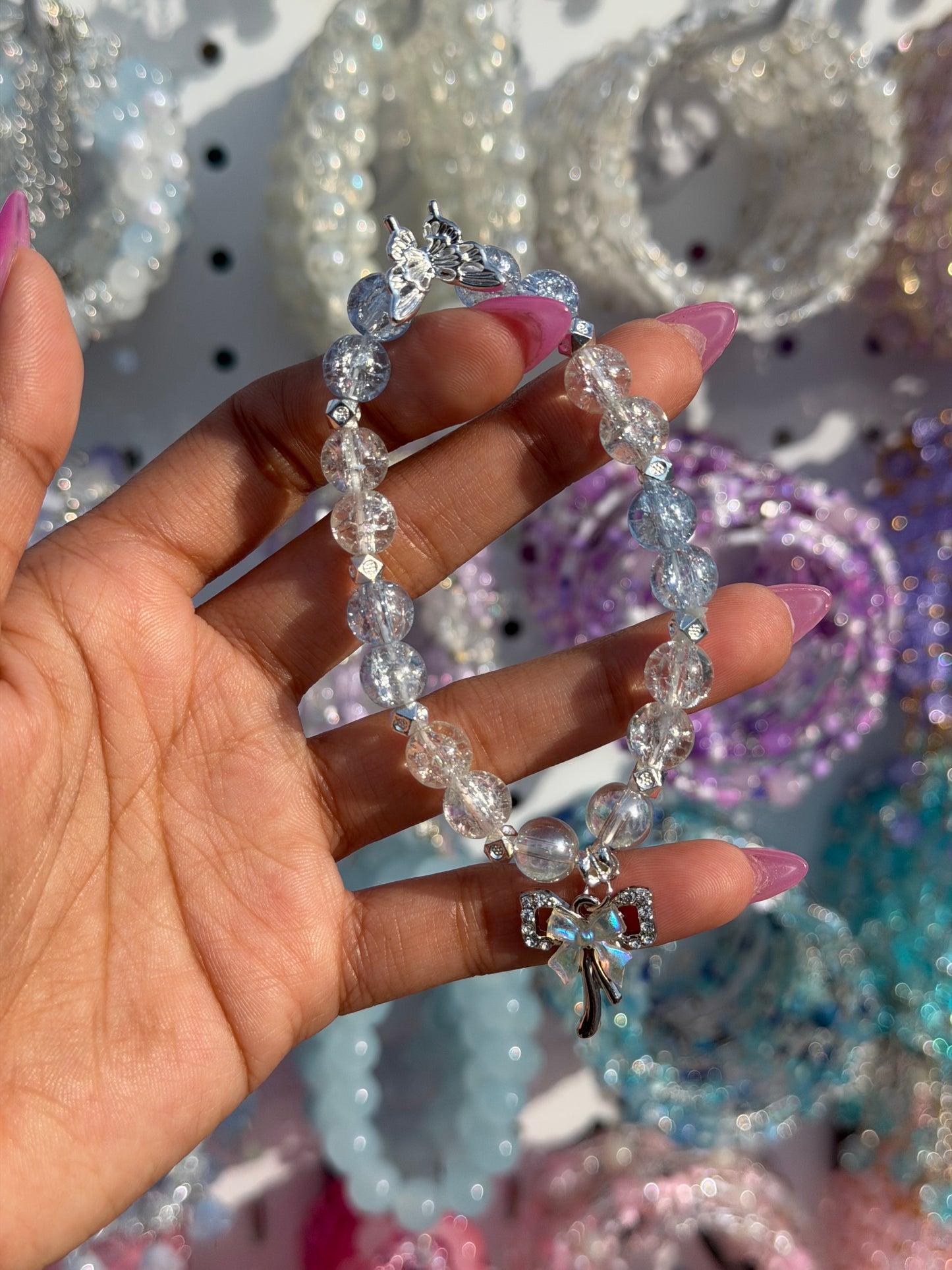 Blue and clear coquette charm beaded bracelet with crackled beads, a butterfly, and a rhinestone bow charm, held by a hand.