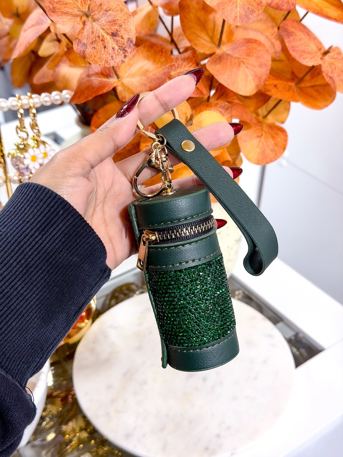 Green glittery keychain with black strap held by a hand against an autumn leaf background