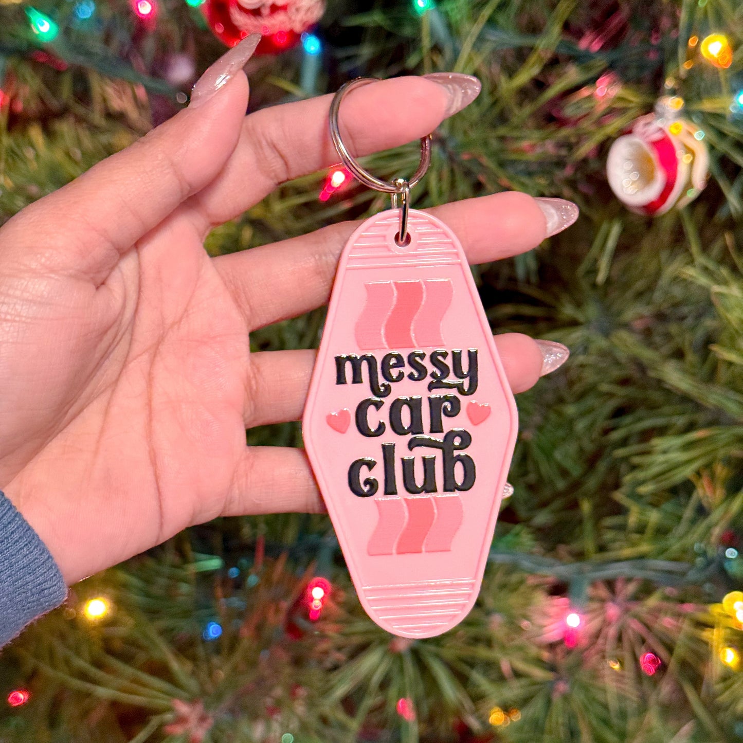 Pink motel keychain with "Messy Car Club" by an unspecified brand, displayed against a Christmas tree.
