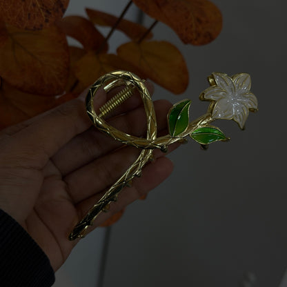 Gold hair claw with a white flower detail and green leaves, designed in a twisted bow shape with a polished metal finish.