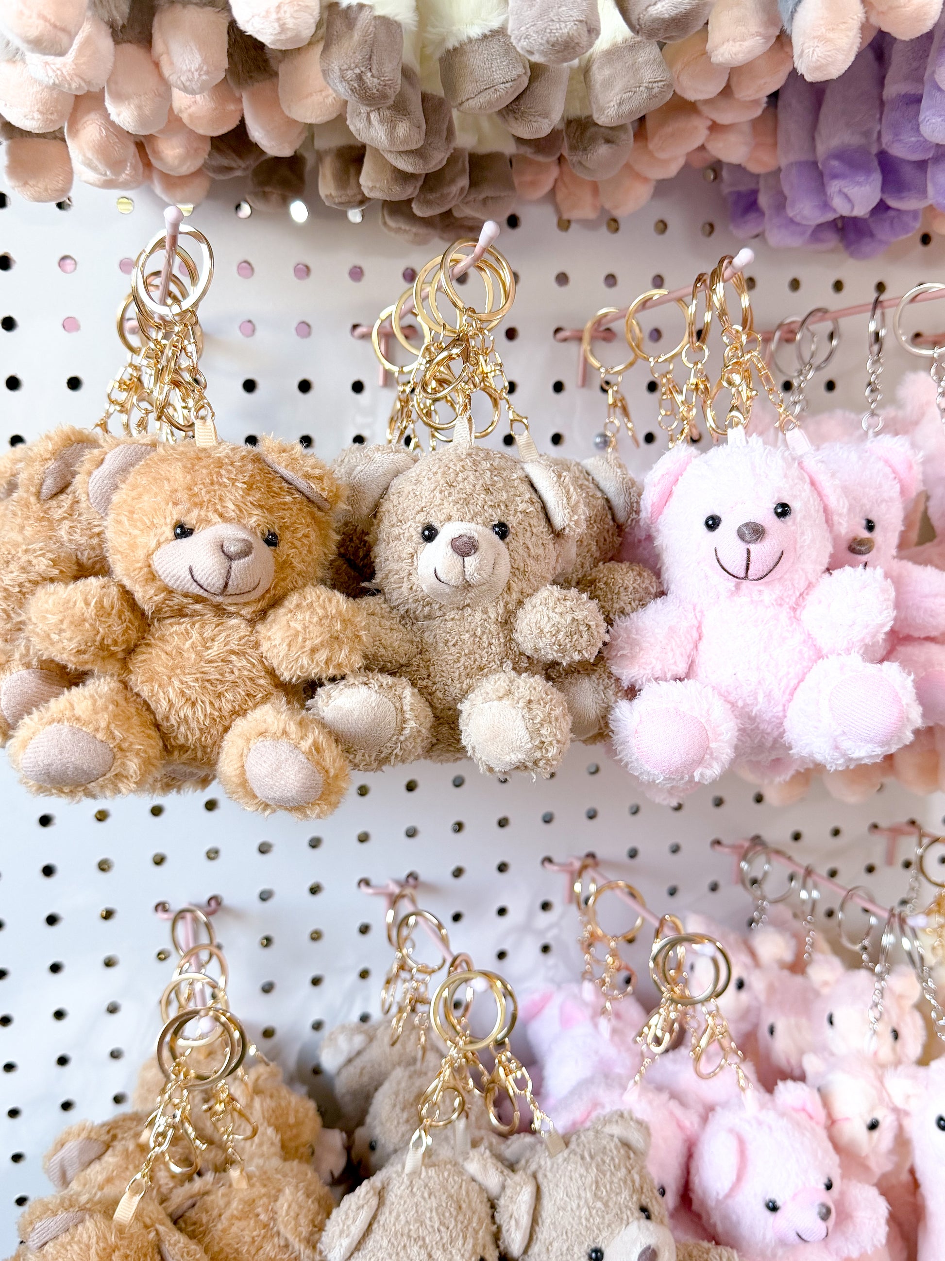 Brown and pink plush cuddle bear bag charms with gold-tone keyrings, displayed on a white pegboard.