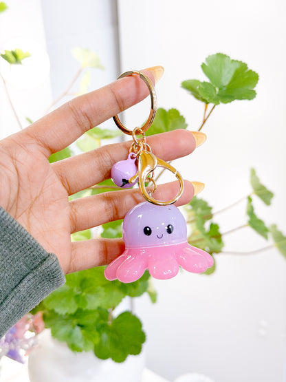Purple and pink Baby Octopus Charm with smiling face and purple bell on a gold keyring, held by a hand.