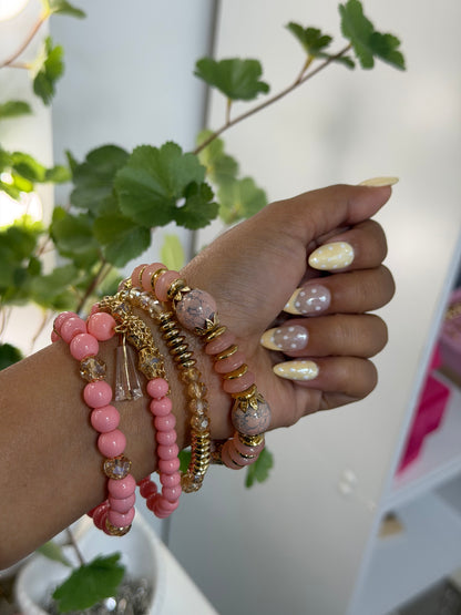 Layered pink and gold beaded charm bracelets worn on a wrist, with a clear cone charm and polka dot nails.