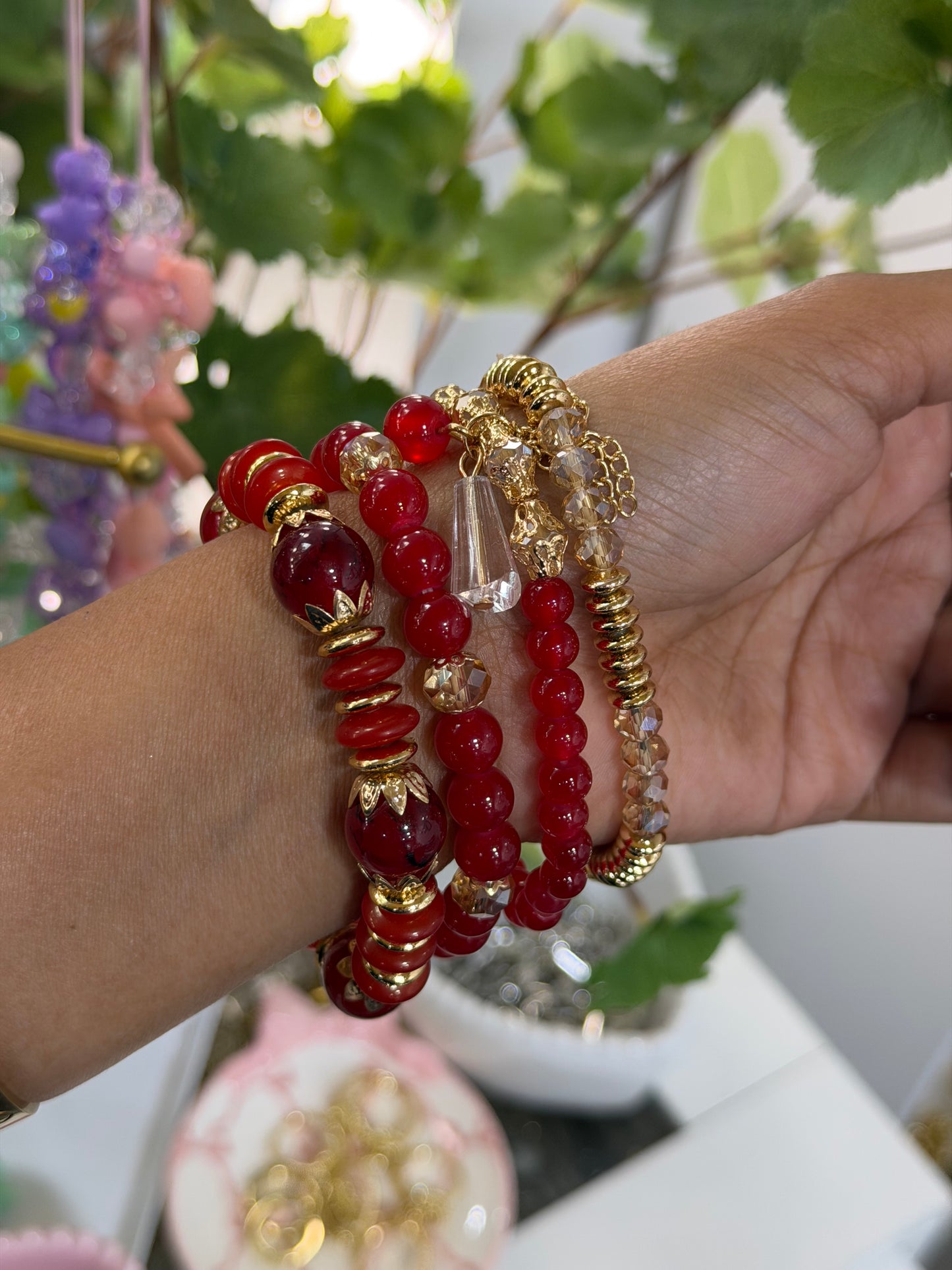 Layered beaded charm bracelets featuring red, gold-tone, and clear faceted beads, worn on a wrist.