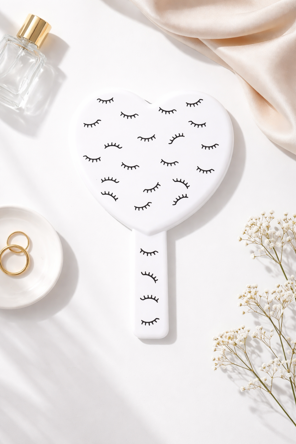White heart-shaped mirror with black eyelash patterns, alongside a perfume bottle, gold rings, cream silk, and white baby's breath flowers.