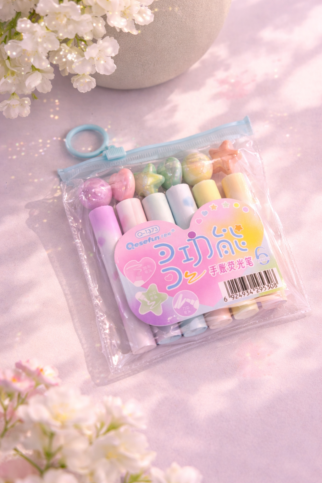 Pastel rainbow highlighter set by Qesefun in a clear pouch, with round, heart, and star shaped caps on a glittery pink surface.