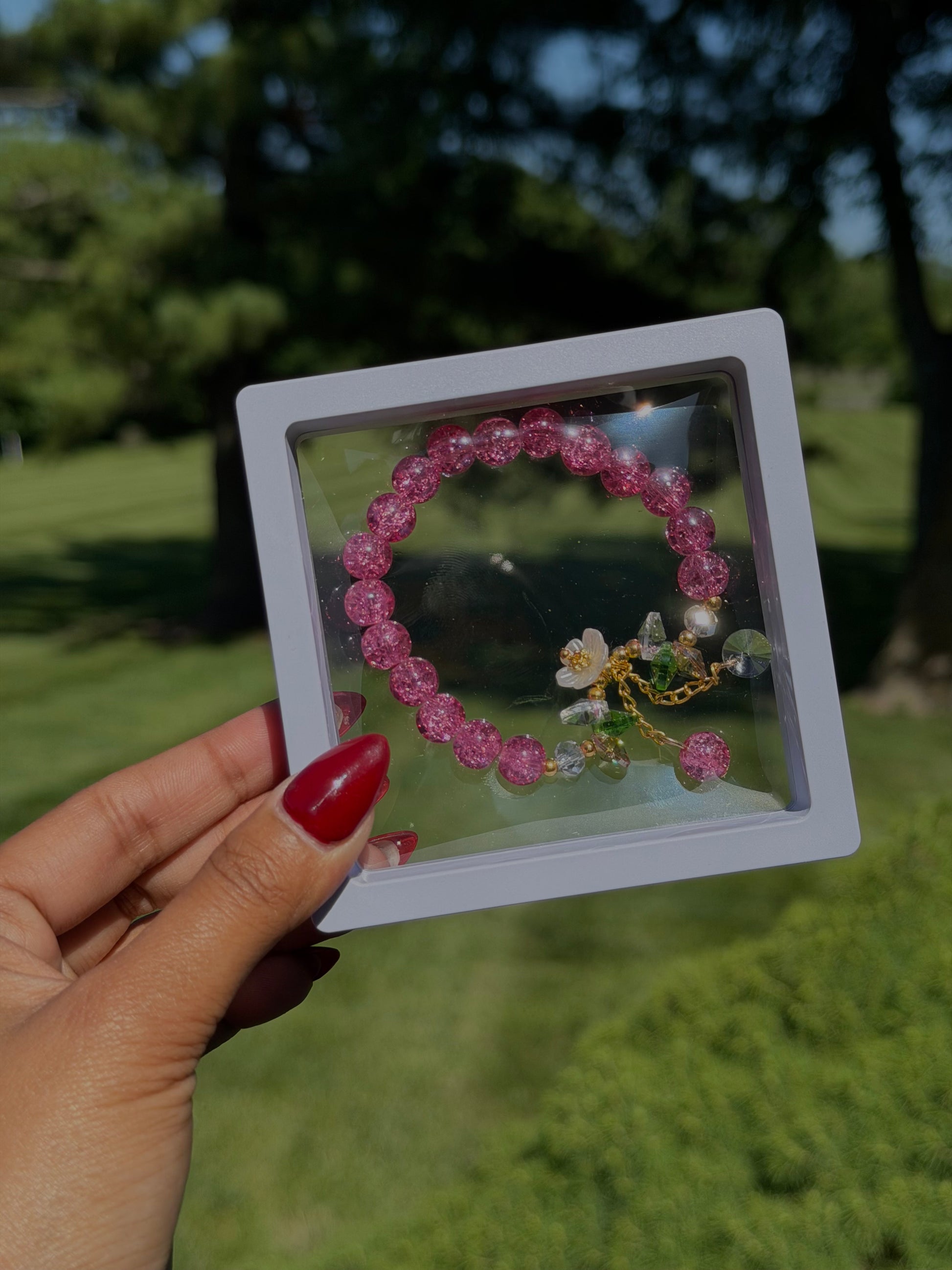 Pink beaded bracelet with floral charm and crystal accents in clear display case, held in hand with red nail polish, outdoor setting