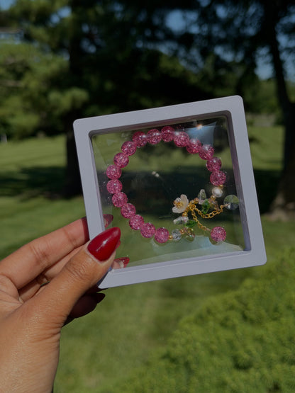 Pink beaded bracelet with floral charm and crystal accents in clear display case, held in hand with red nail polish, outdoor setting