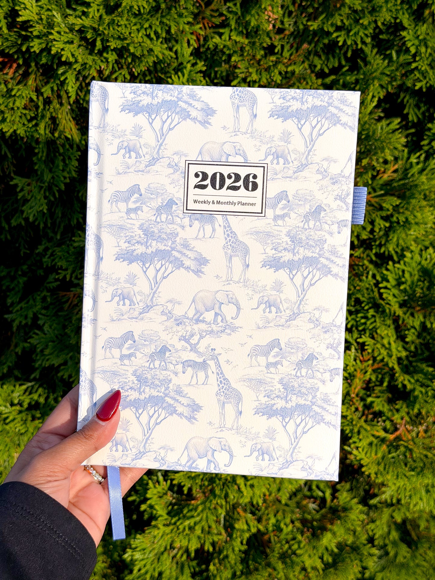 White 2026 weekly & monthly planner with blue safari animal pattern, held in hand over green foliage, featuring ribbon bookmark and textured cover.