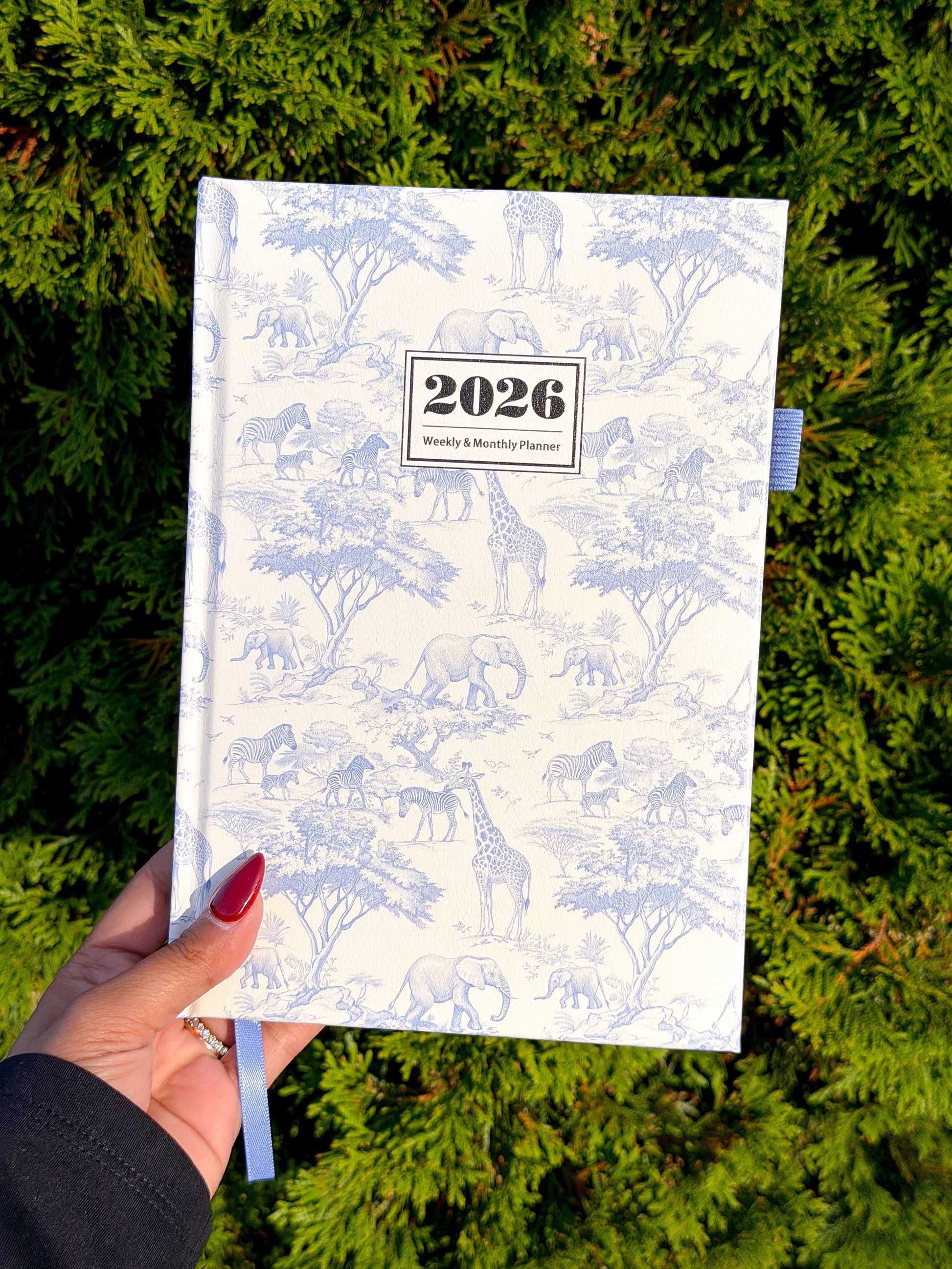 White 2026 weekly & monthly planner with blue safari animal pattern, held in hand over green foliage, featuring ribbon bookmark and textured cover.