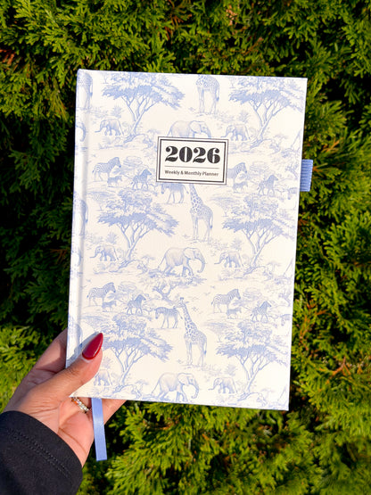 White 2026 weekly & monthly planner with blue safari animal pattern, held in hand over green foliage, featuring ribbon bookmark and textured cover.