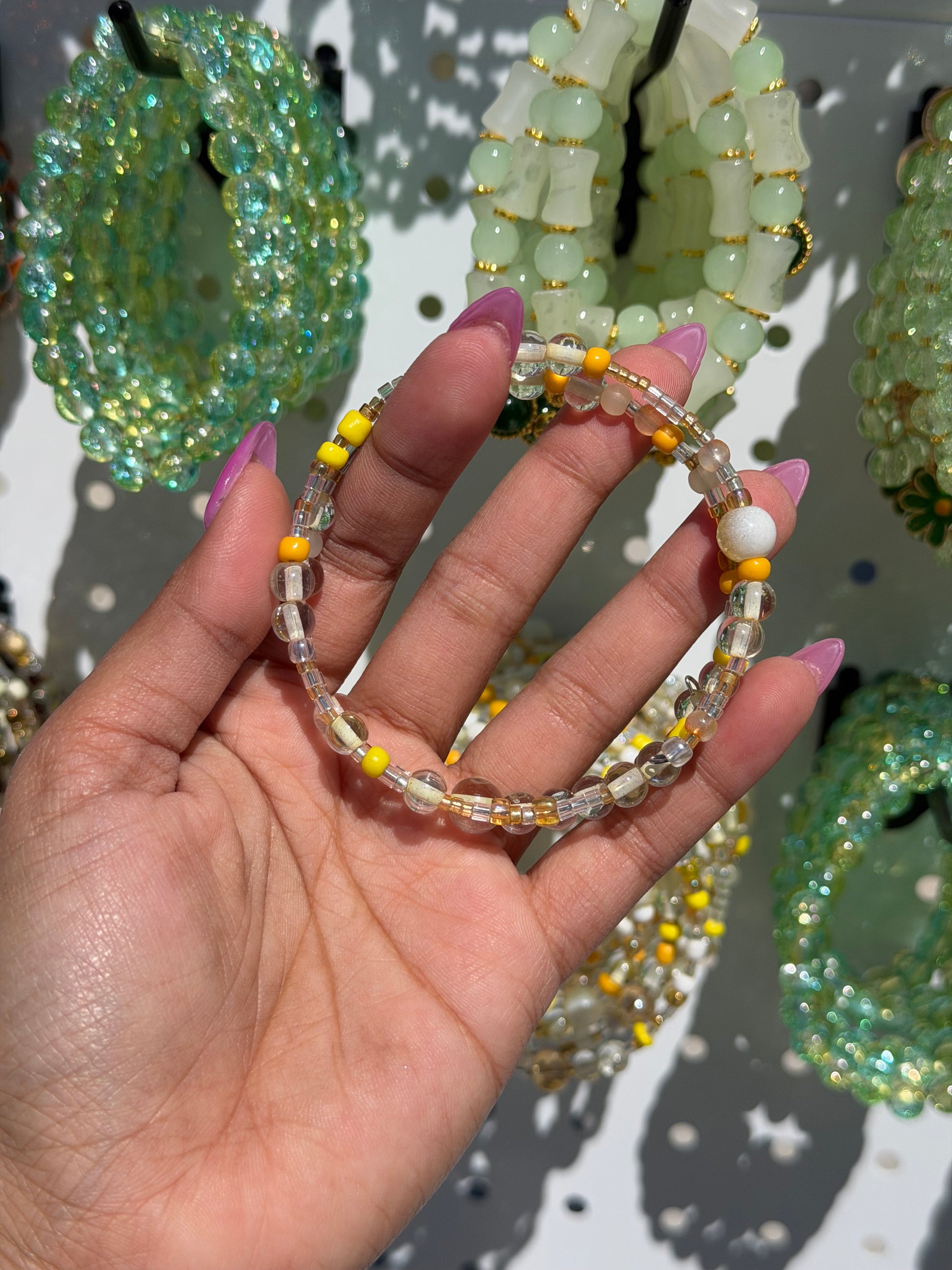 Layered Beaded Bracelets with clear, yellow, and white beads, held by a hand; blurred green and white bracelets in background.