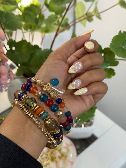 Multi-colored layered beaded charm bracelets on a wrist with yellow polka dot nails, set against blurred greenery.