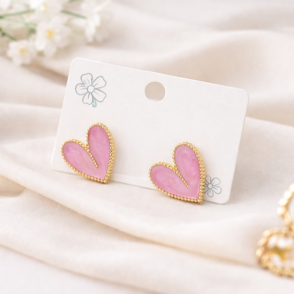 Marbled pink heart stud earrings with a gold beaded border, displayed on a white card against soft white fabric.