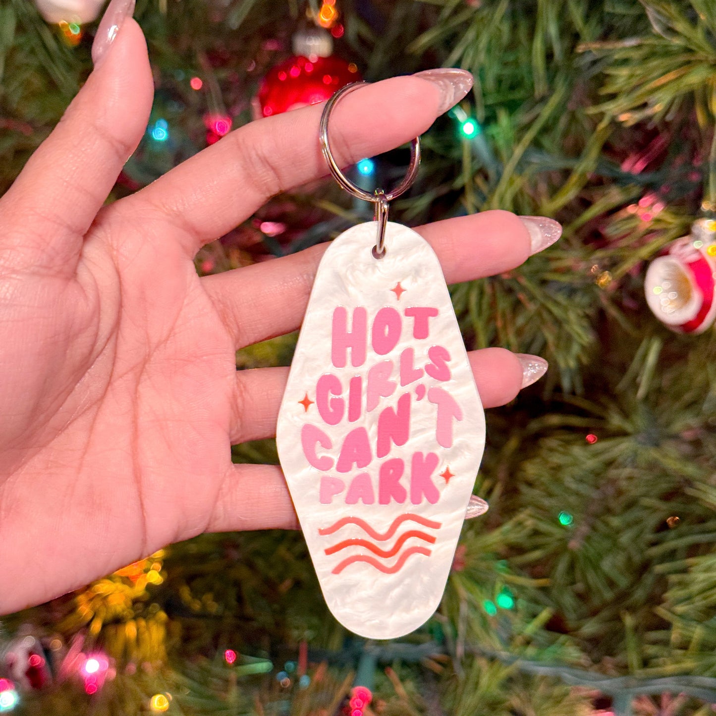 Motel keychain by an unknown brand with a "Hot Girls Can't Park" slogan in pink, held in hand against a Christmas tree.
