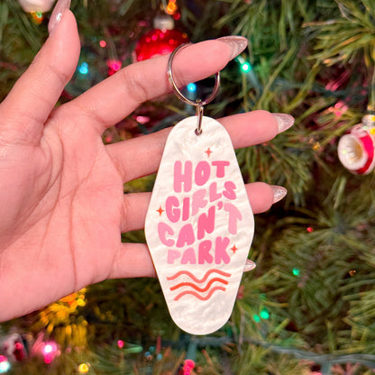 Motel keychain by an unknown brand with a "Hot Girls Can't Park" slogan in pink, held in hand against a Christmas tree.
