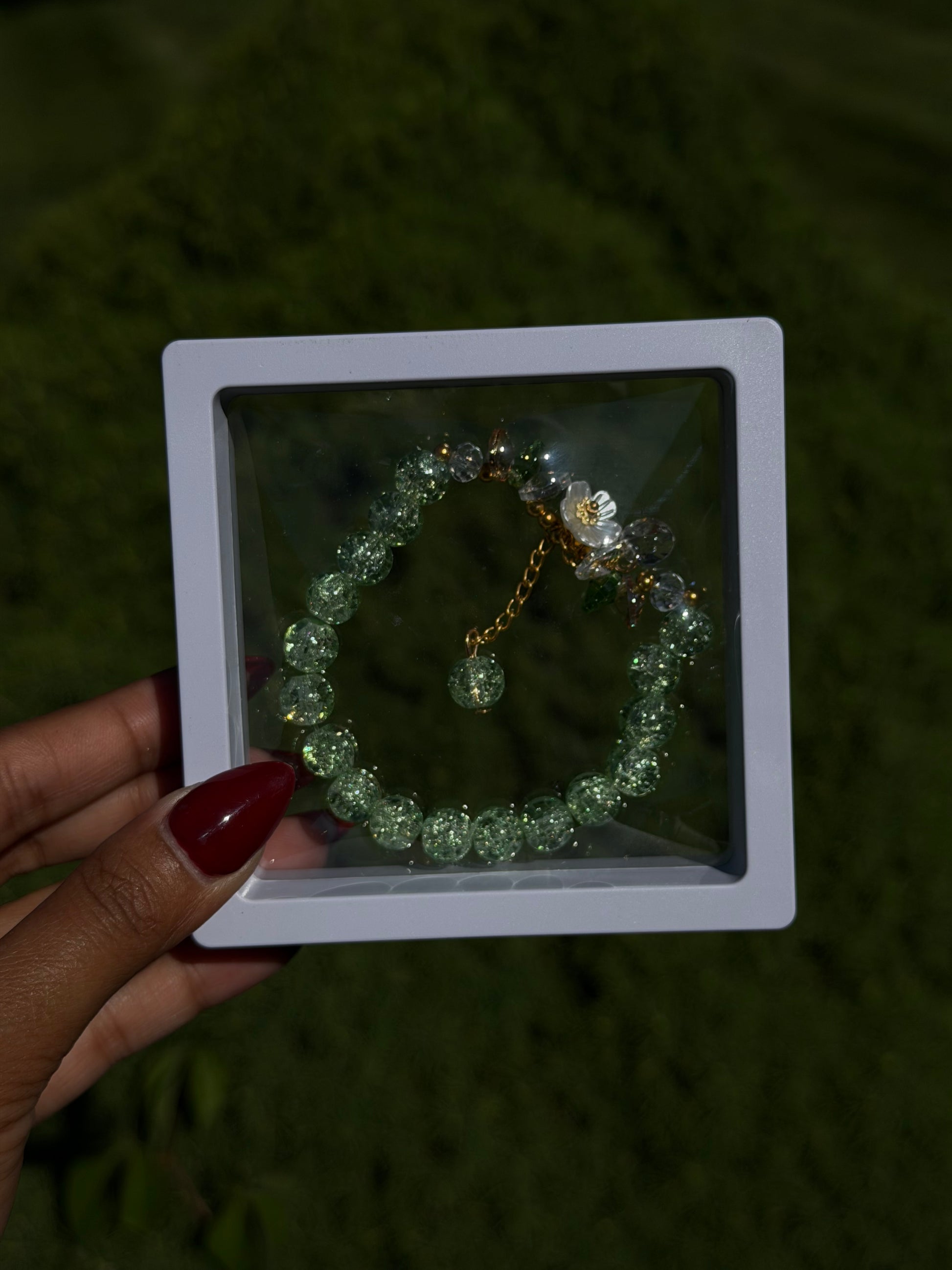 Green beaded bracelet with glittery texture and floral charm in clear case, held in hand with red nail polish, outdoor grass background