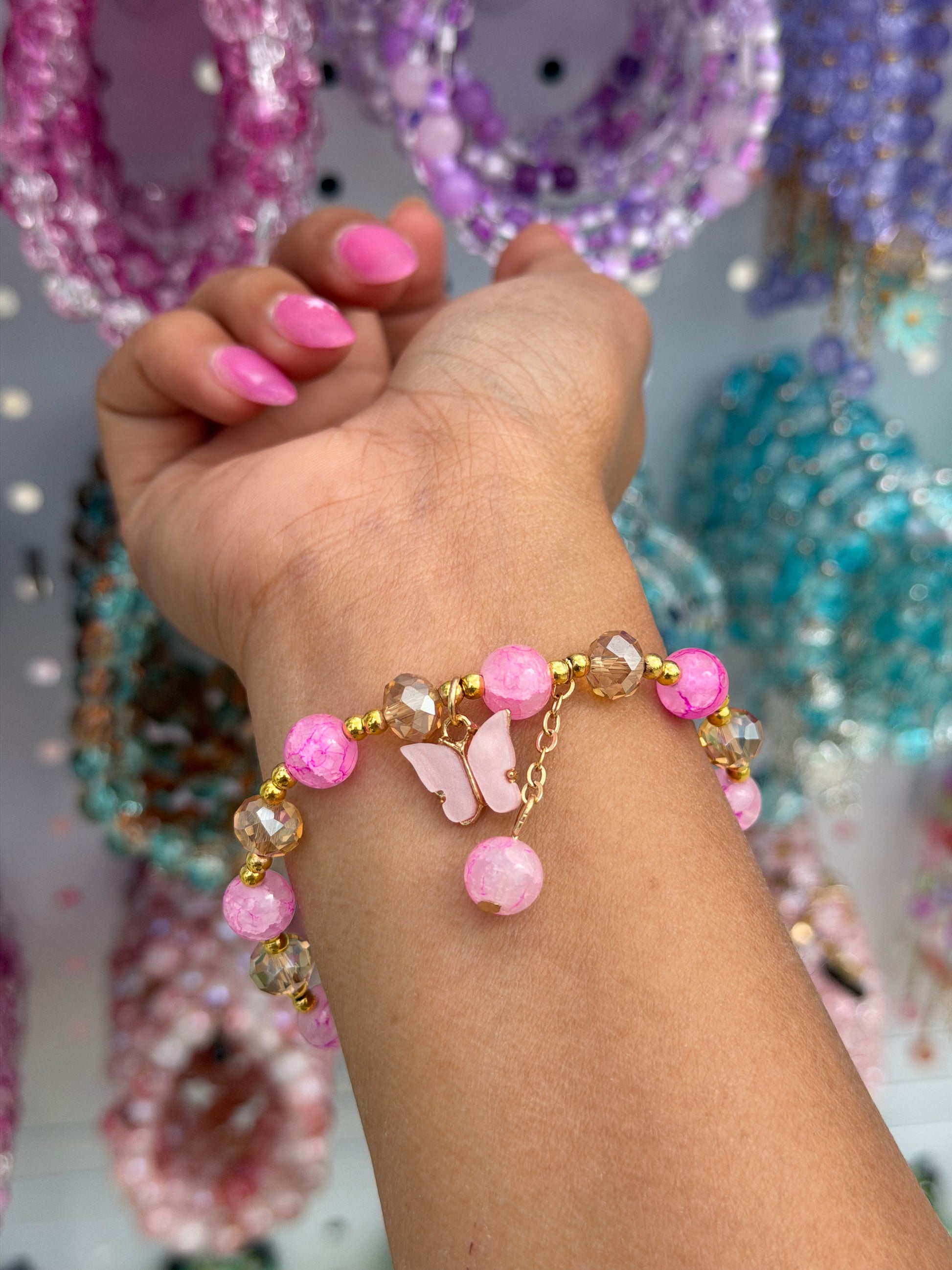 Pink crystal butterfly beaded bracelet with crackled pink and golden faceted beads, a pink butterfly charm, and a dangling pink bead, worn on a hand with bright pink nails.