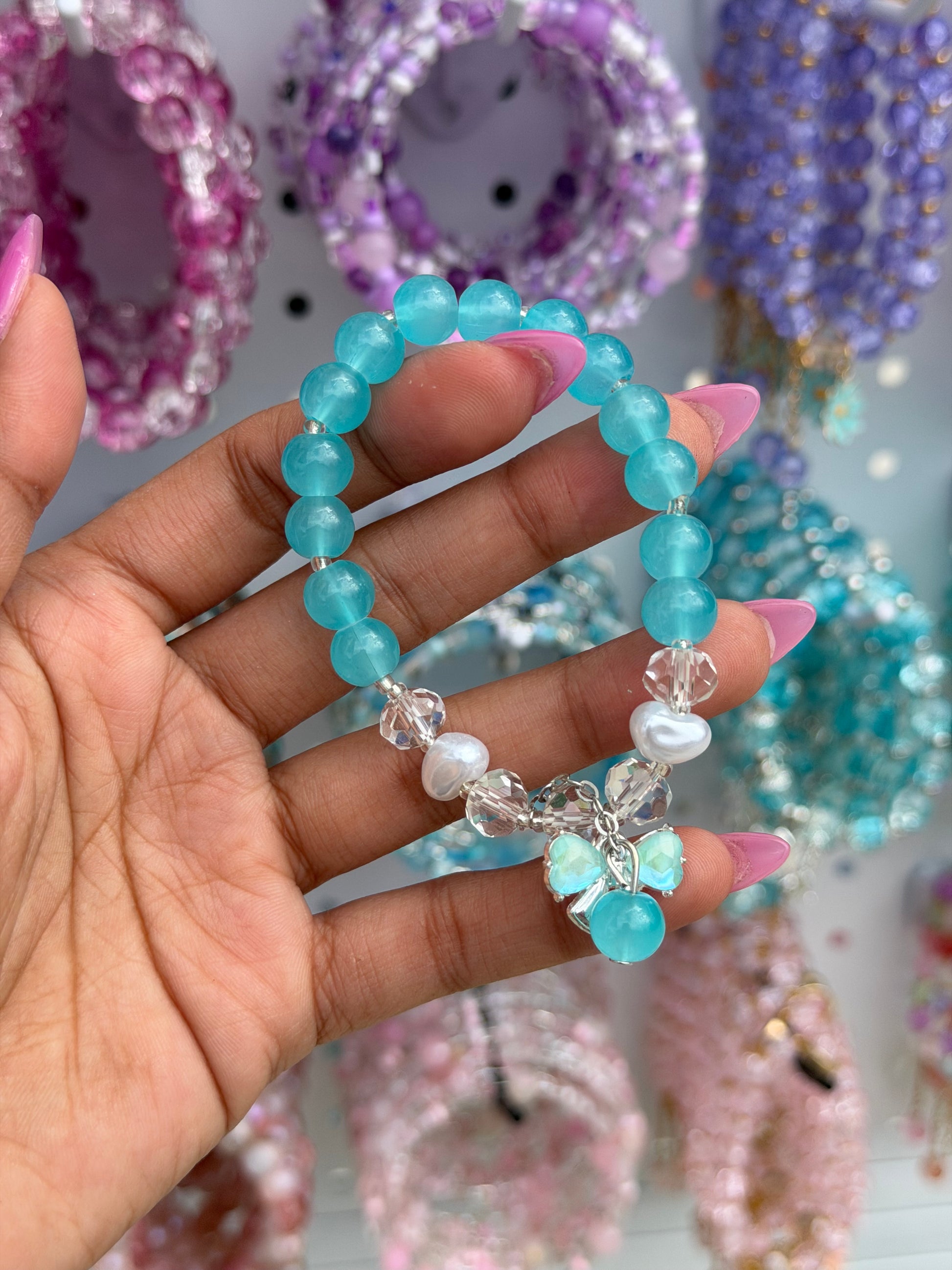 Blue beaded bracelet with clear crystal spacers, a heart-shaped pearl, and a blue bow charm, held by a pink-nailed hand.