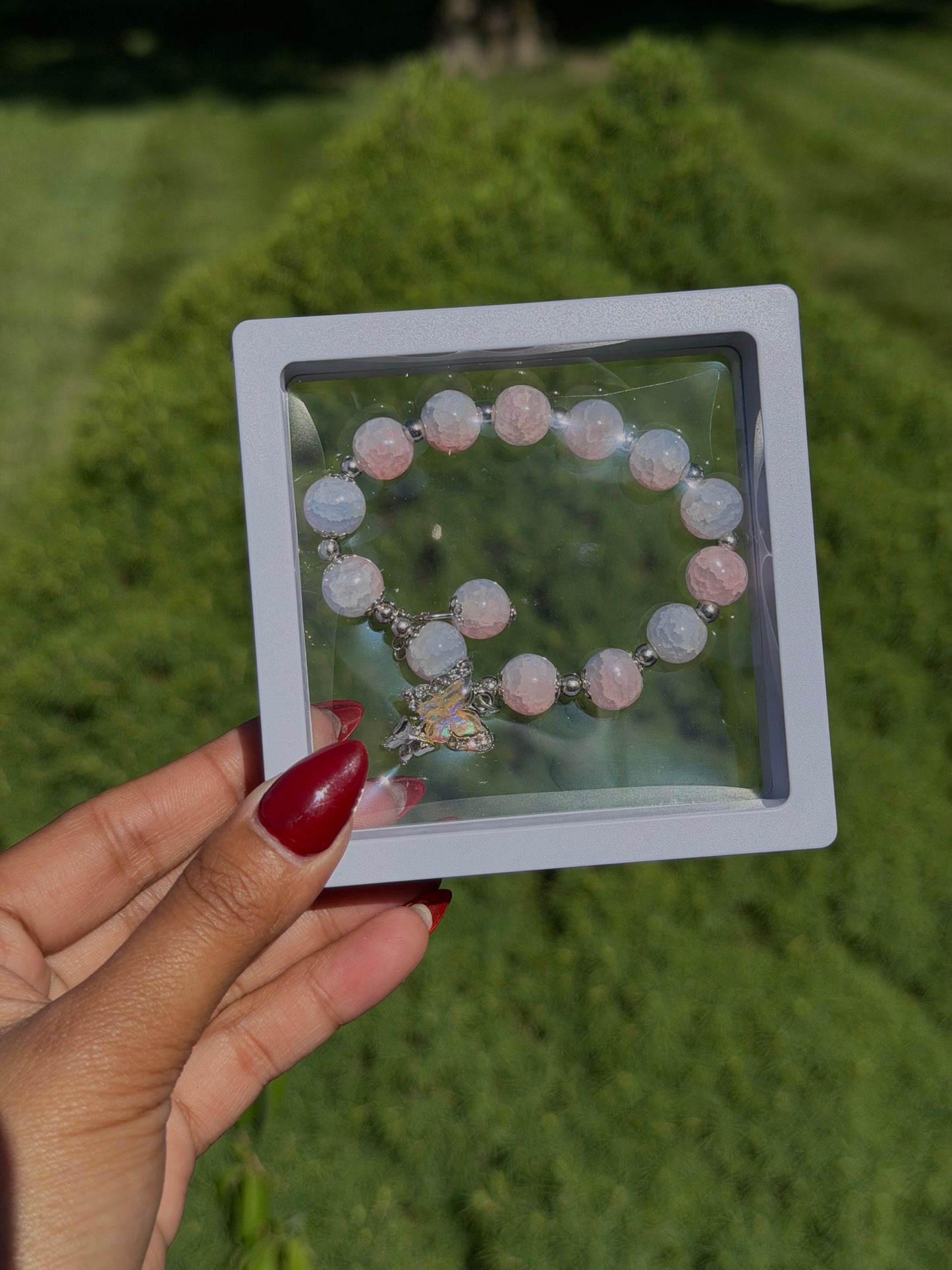 Pink and white beaded bracelet with iridescent butterfly charm in clear display case, held in hand with red nails, outdoor green background