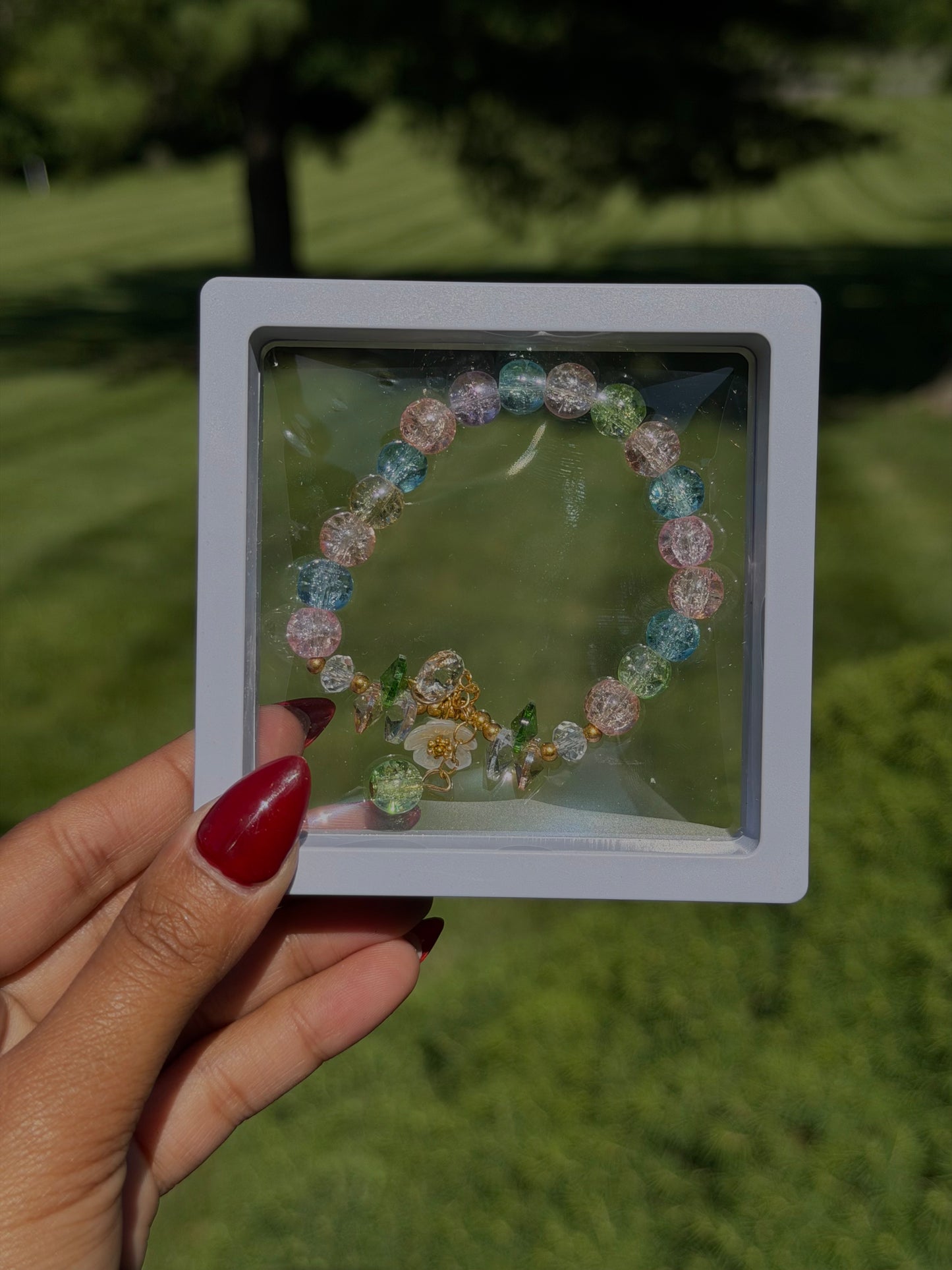 Colorful beaded bracelet with iridescent glass beads and gold-tone charms in clear display case, held by hand with red nail polish, outdoors.