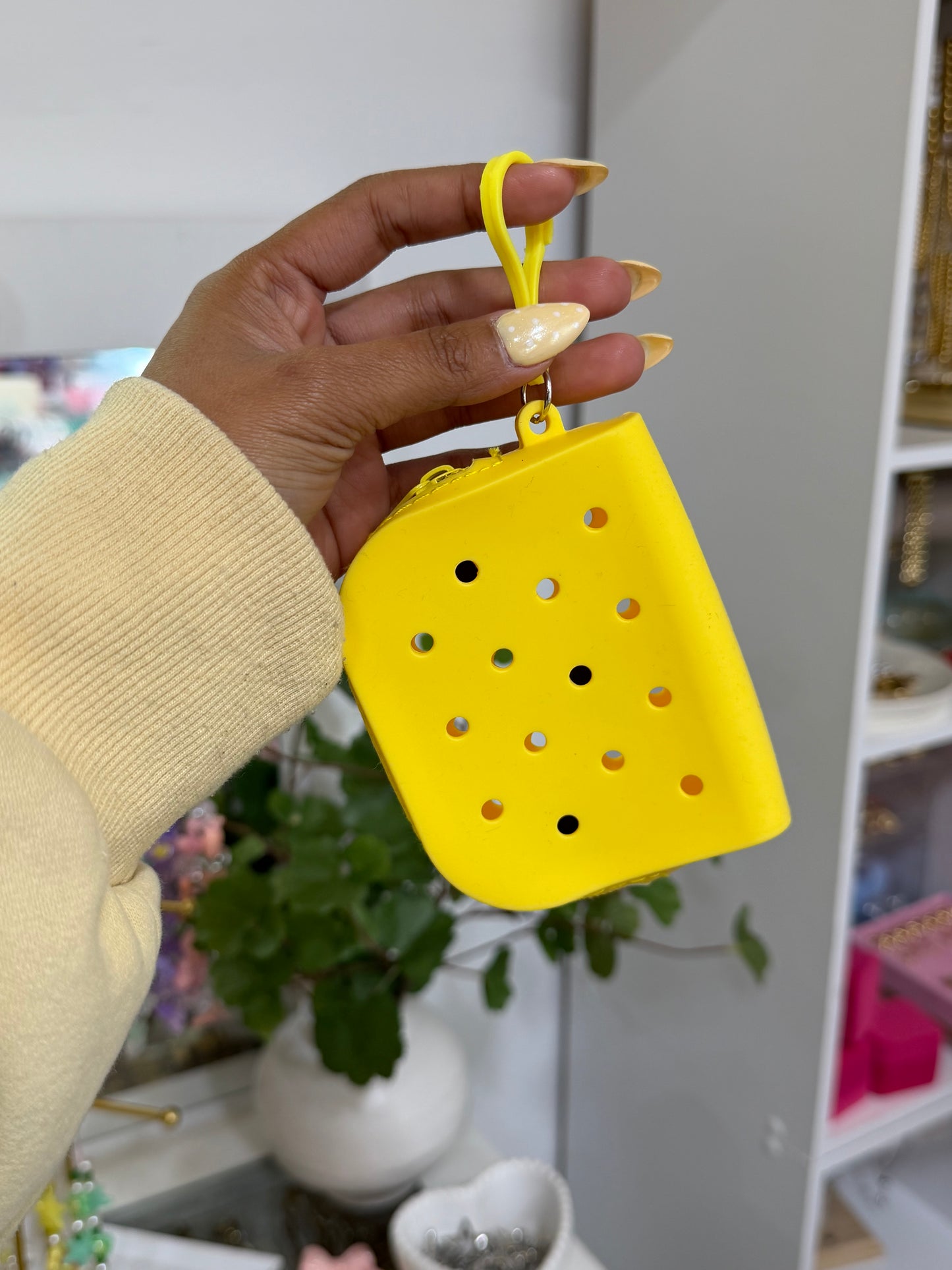 Triangular yellow silicone charm pouch with holes and a loop, held by a hand in a yellow sweater.