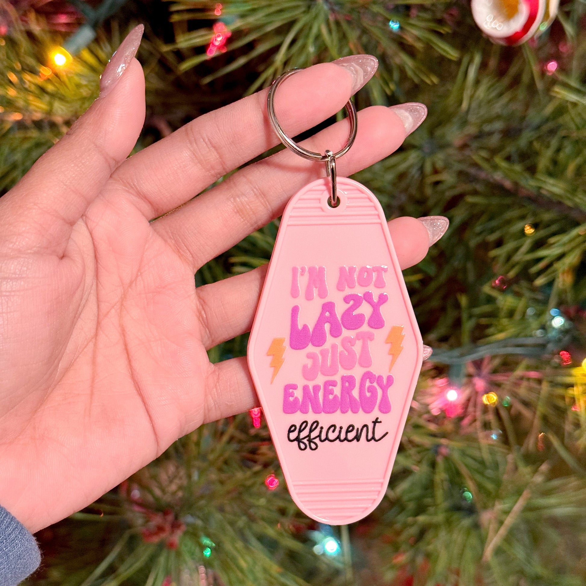 Pink motel keychain with a funny saying by vendor, held in someone's hand with a blurred Christmas tree background.
