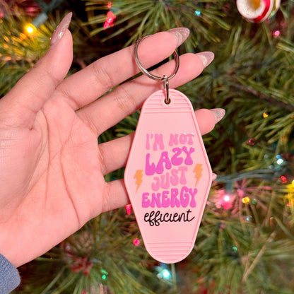 Pink motel keychain with a funny saying by vendor, held in someone's hand with a blurred Christmas tree background.
