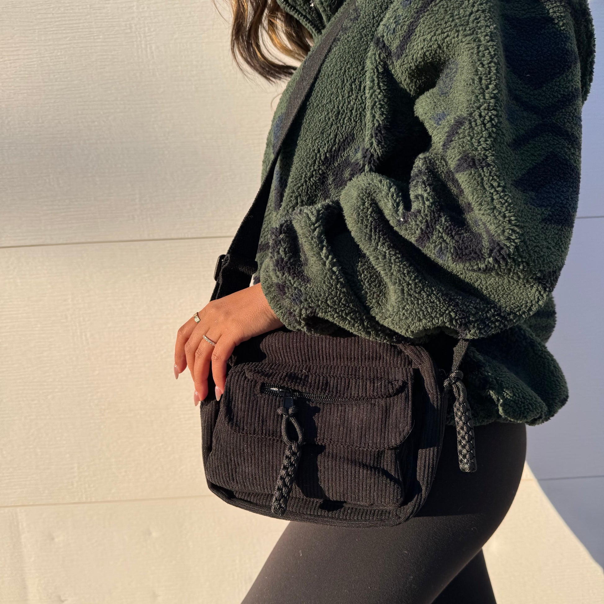Black corduroy crossbody bag with braided zipper pulls, paired with a green patterned sherpa jacket and leggings.