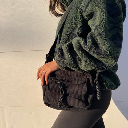 Black corduroy crossbody bag with braided zipper pulls, paired with a green patterned sherpa jacket and leggings.