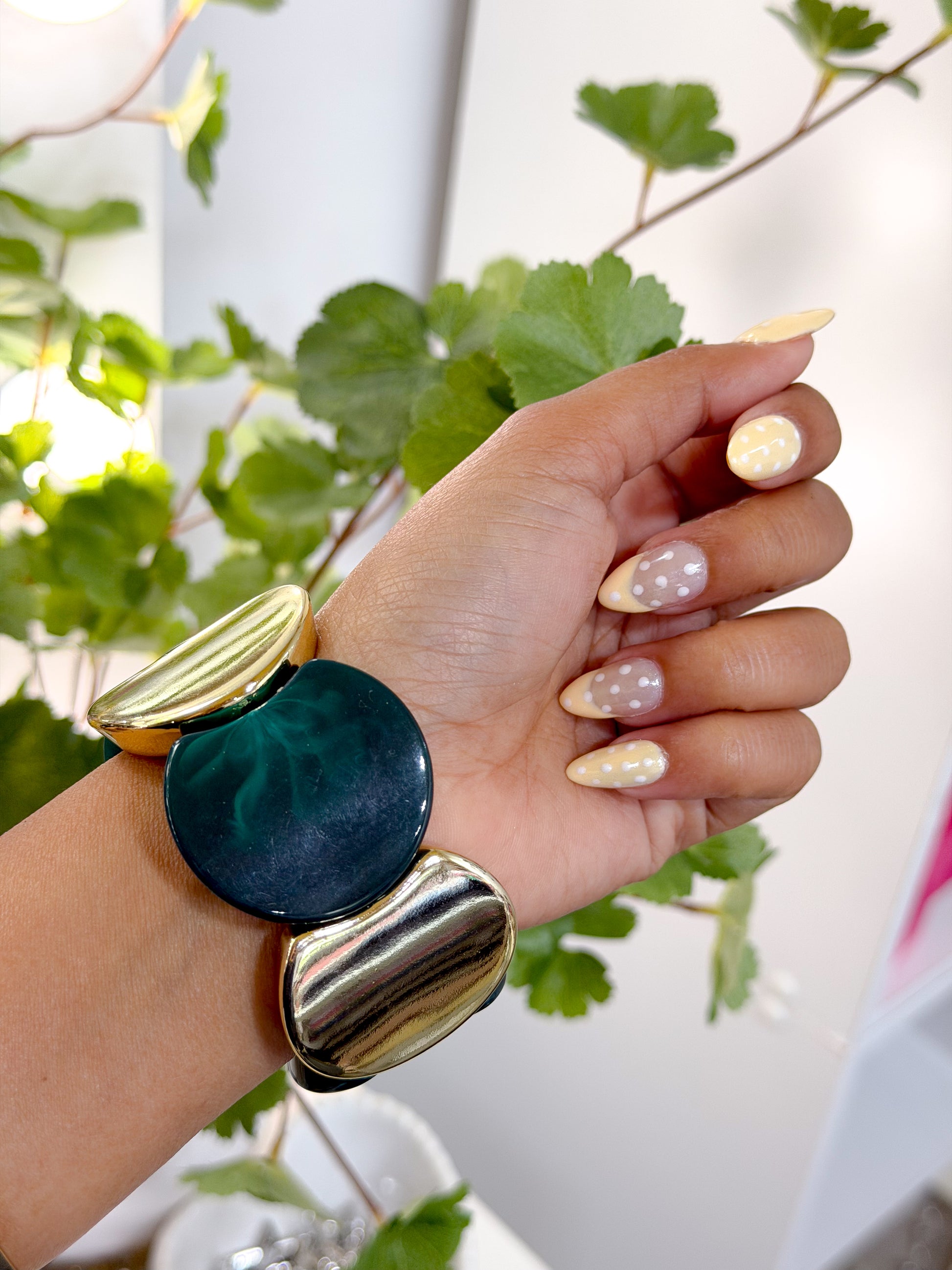 Chunky statement bracelet in green and gold, worn on a wrist with yellow and nude polka dot nails, surrounded by green leaves.