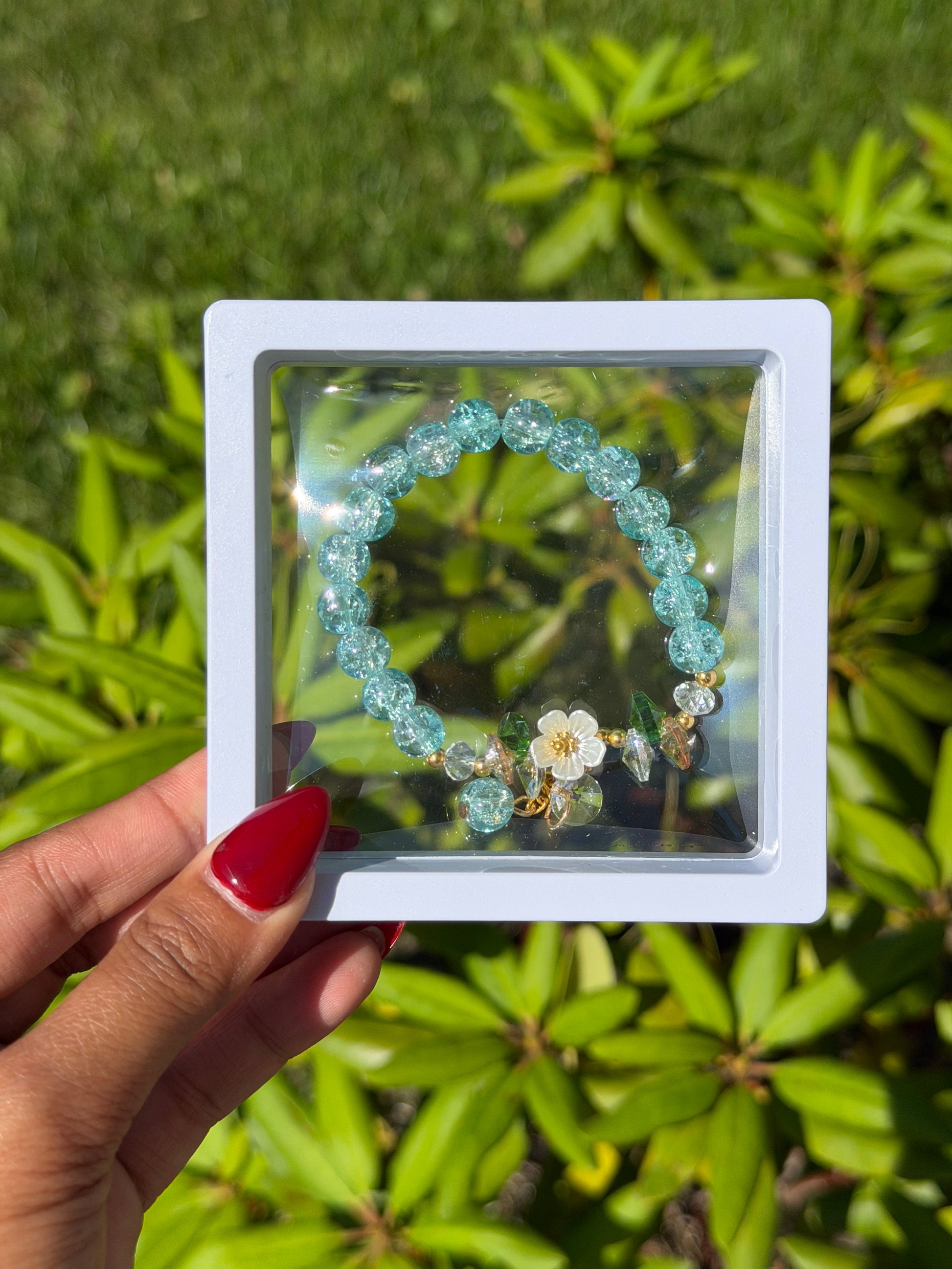 Turquoise beaded bracelet with white flower charm and crystal accents in clear display case, held against green foliage background