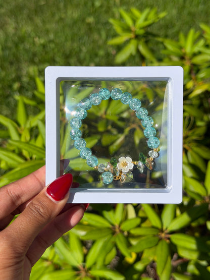 Turquoise beaded bracelet with white flower charm and crystal accents in clear display case, held against green foliage background
