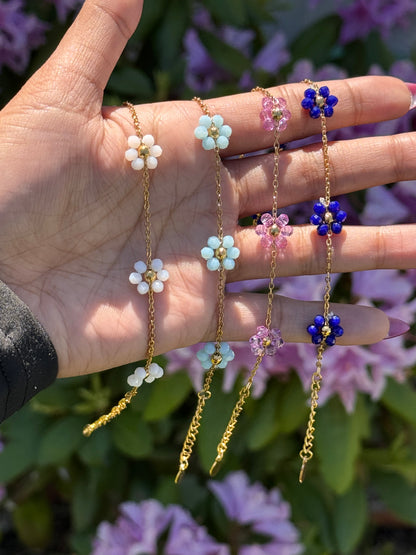 Four gold Mini Daisy Bracelets with white, light blue, pink, and dark blue beaded flowers, dangling from a hand against blurred purple flowers.