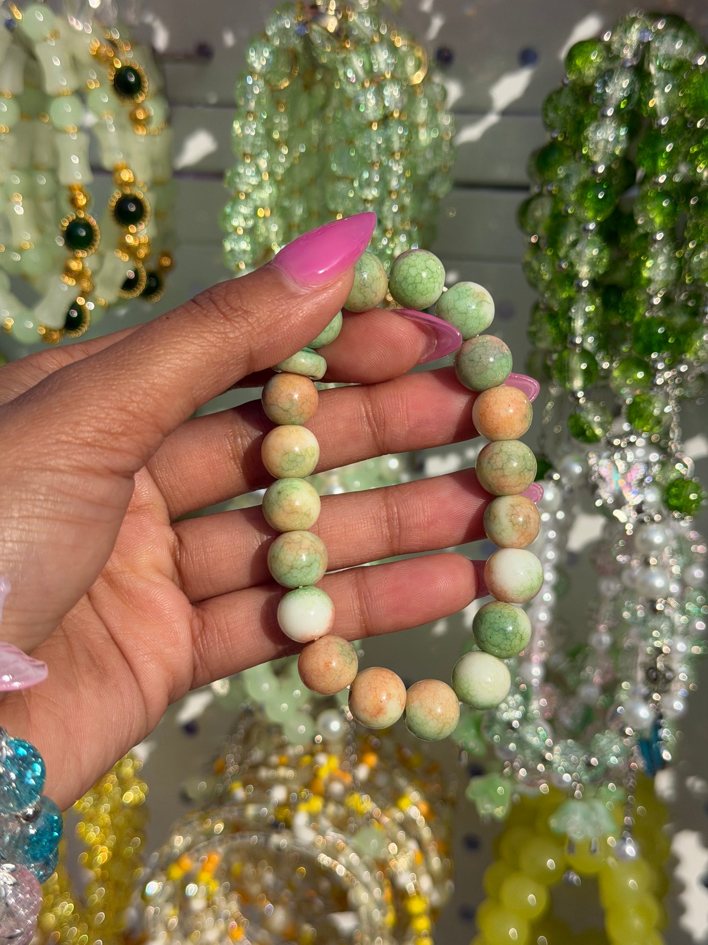 Green and peach marble beaded bracelet held by a hand with pink manicured nails, against a blurred background of colorful jewelry.