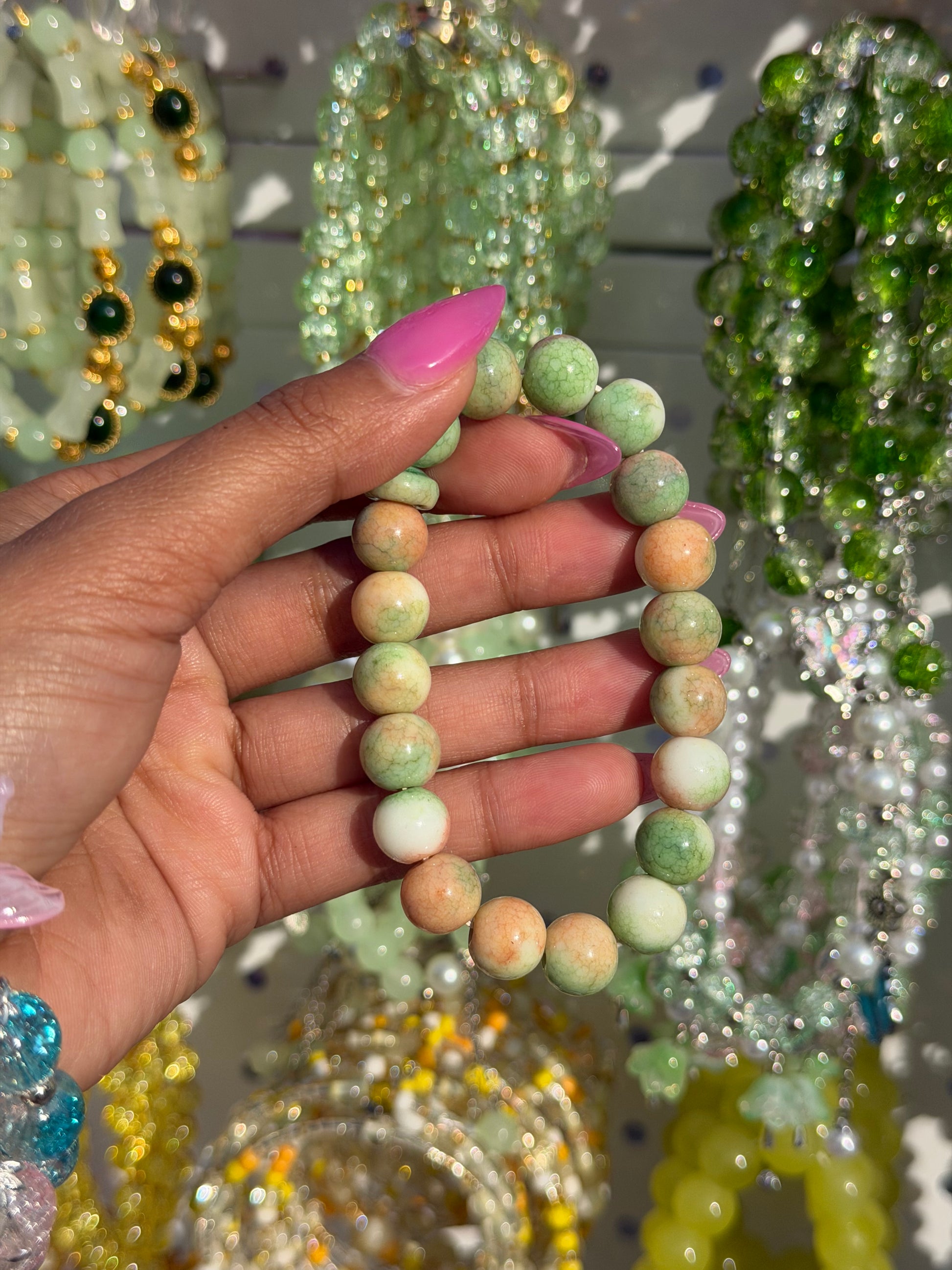 Green and peach marble beaded bracelet held by a hand with pink manicured nails, against a blurred background of colorful jewelry.